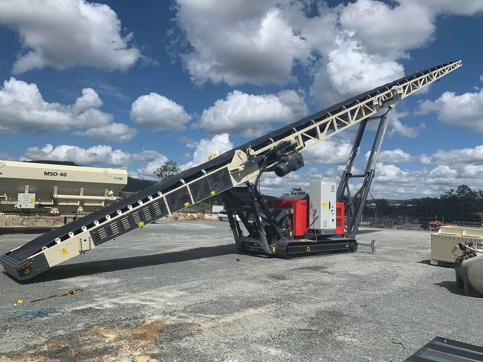 A mobile conveyor belt system outdoors on a gravel surface with a blue sky and clouds in the background.