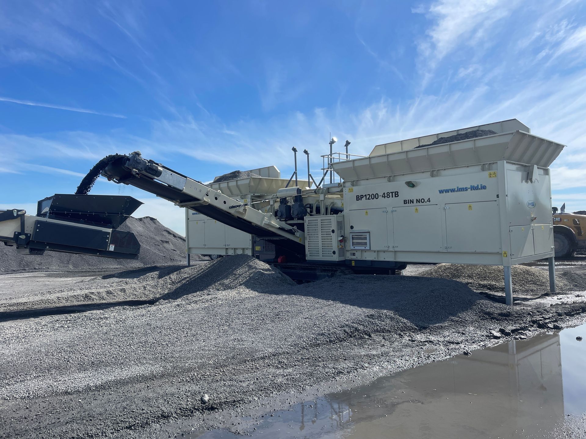 A large white industrial machine, likely a rock crusher, processing gravel on a sunny day with a blue sky.