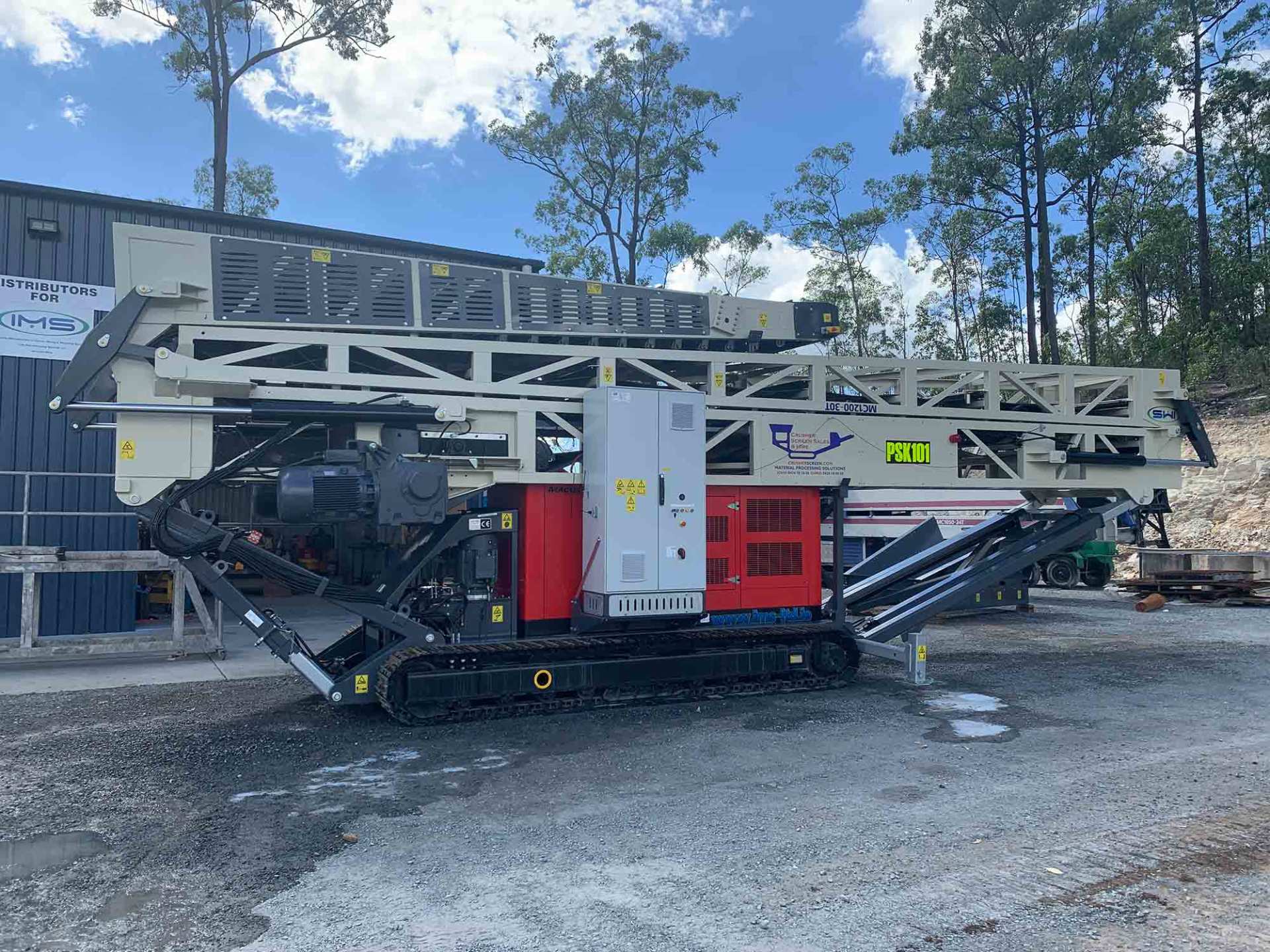 A large industrial machine with a red and white chassis and black tracks sits outdoors, likely a rock crusher or similar equipment.