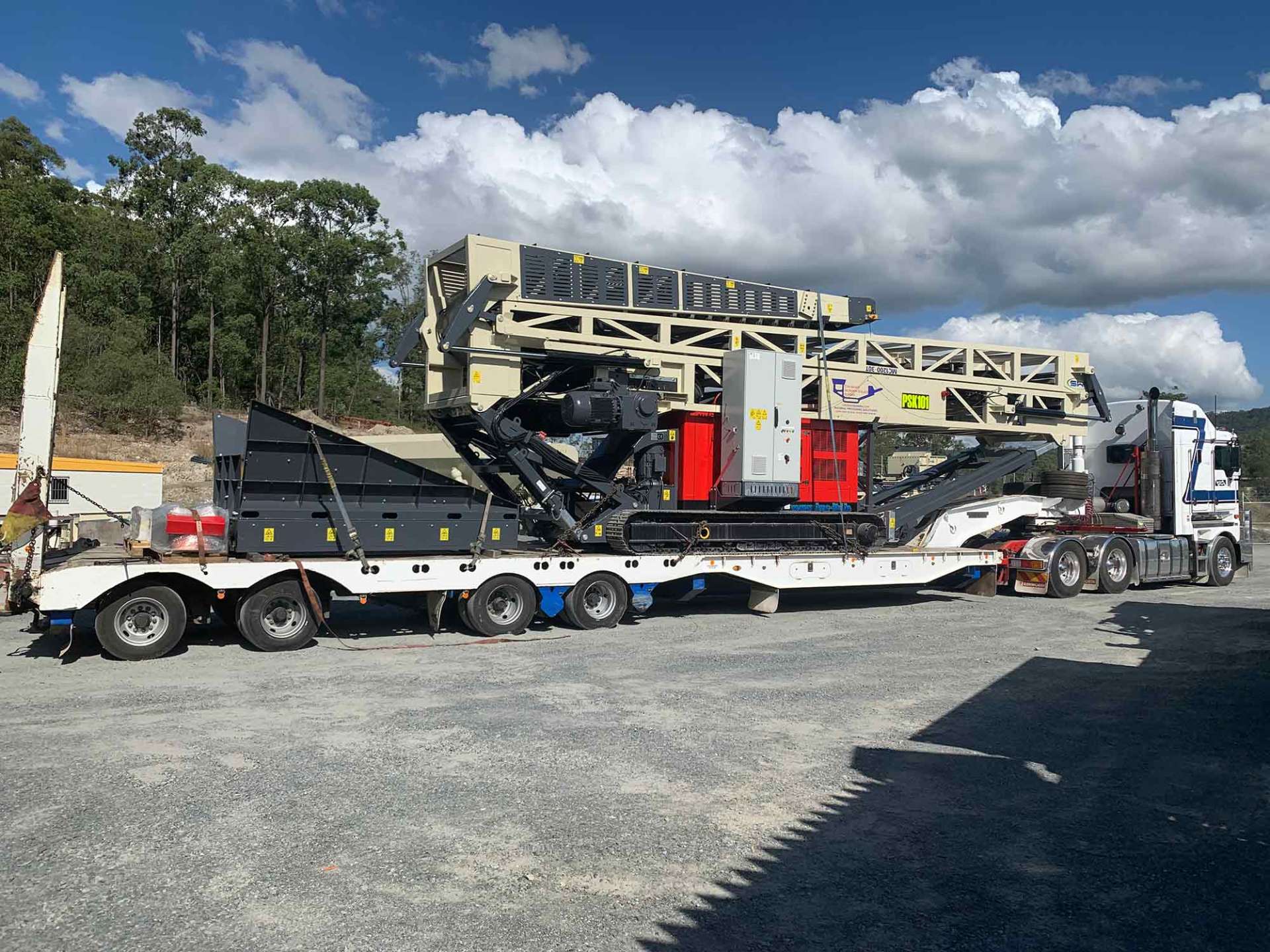 Heavy machinery transported on a flatbed truck. The equipment includes a crusher, painted gray, red, and white. The setting is outdoors with a blue sky and trees in the background.