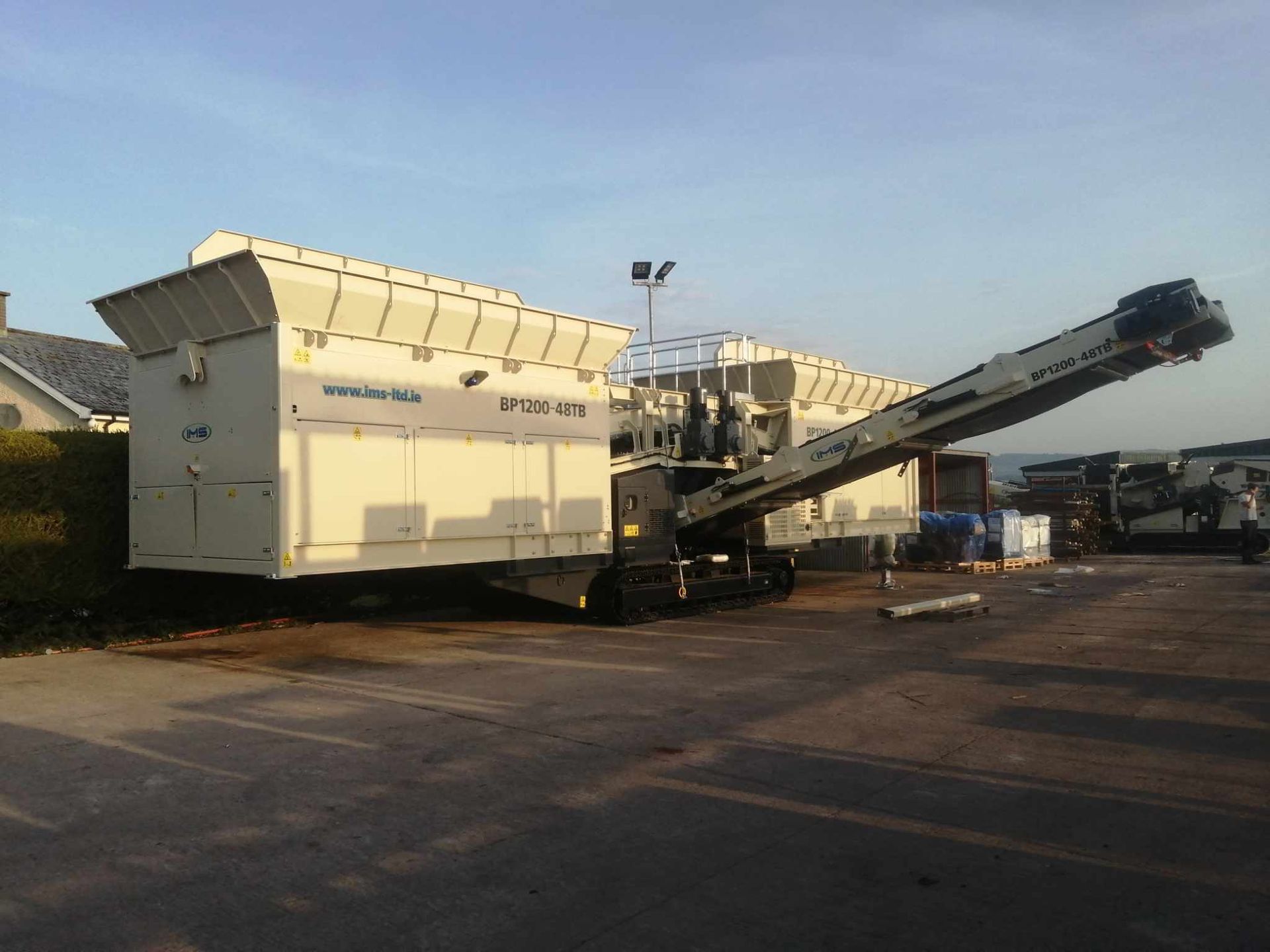 A large, white mobile crushing machine on tracks in a parking lot, with a conveyor belt extending upwards.