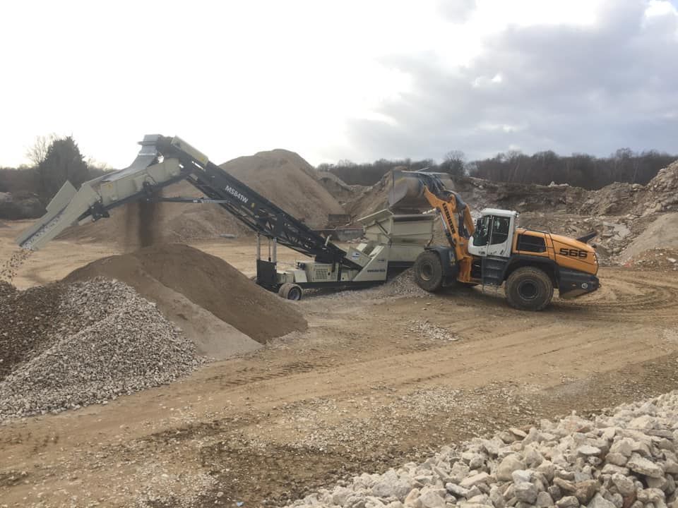 A red and black rock crusher working outdoors, producing piles of crushed stone under a blue sky.