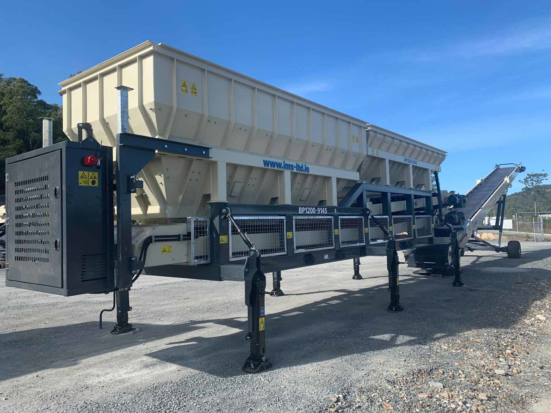 A large, industrial machine for processing materials, likely gravel or concrete, outdoors under a blue sky. The machine is beige and black.