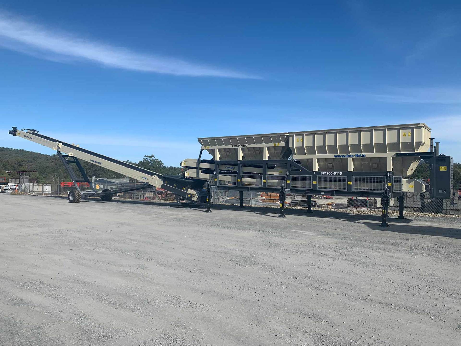 Mobile crushing plant with conveyor belt, on gravel under a blue sky.