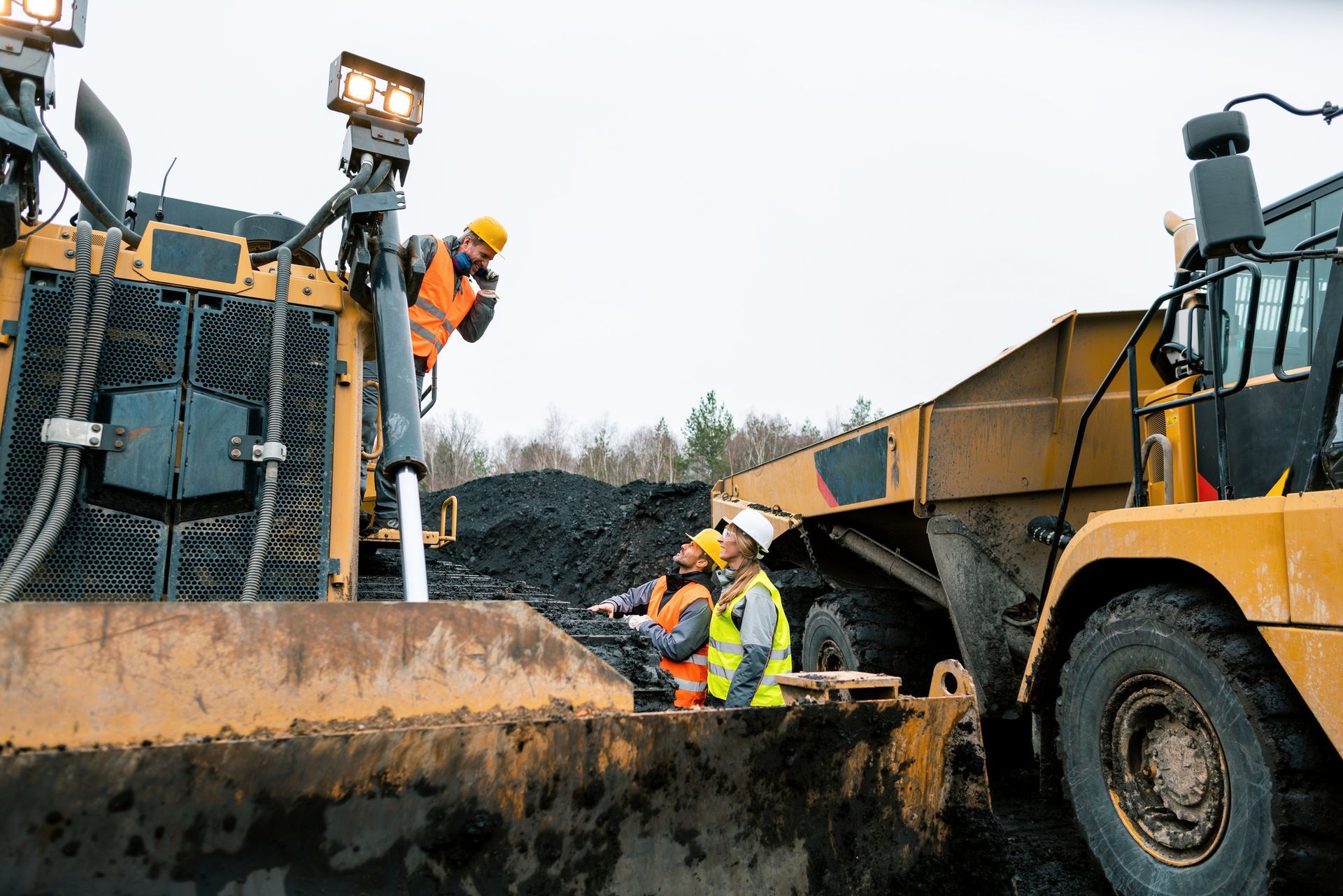Workers in safety vests and hard hats inspecting a large yellow mining vehicle loaded with black material.