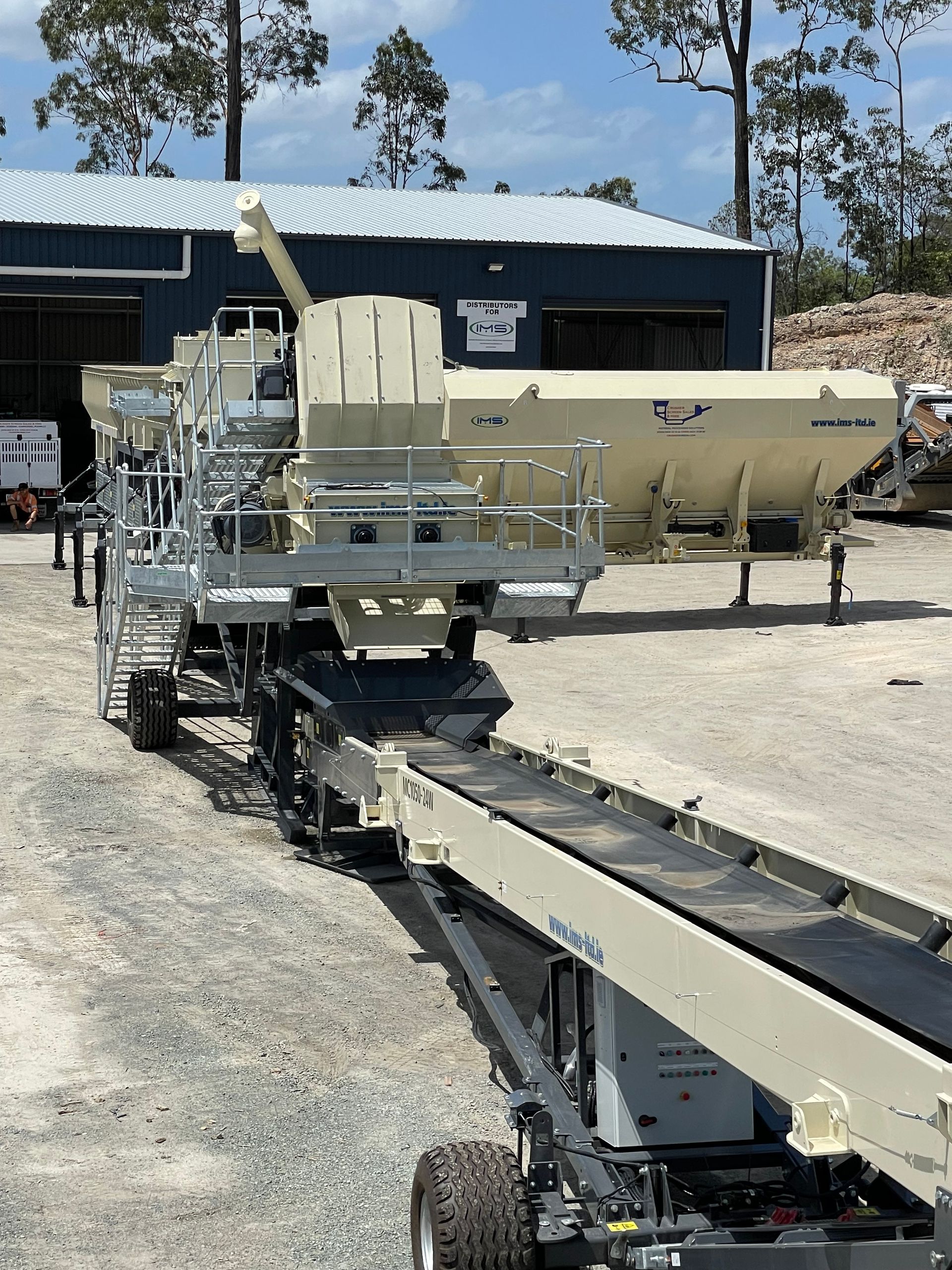A beige industrial machine with conveyor belts, likely used for construction, outdoors in front of a warehouse on a sunny day.