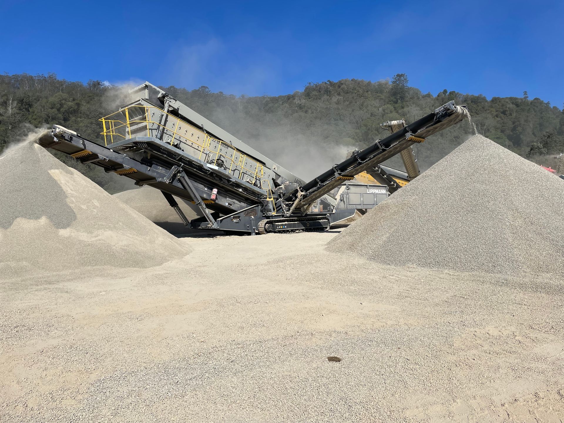 Rock-crushing machine separating gravel into piles at a quarry, with trees and blue sky in the background.