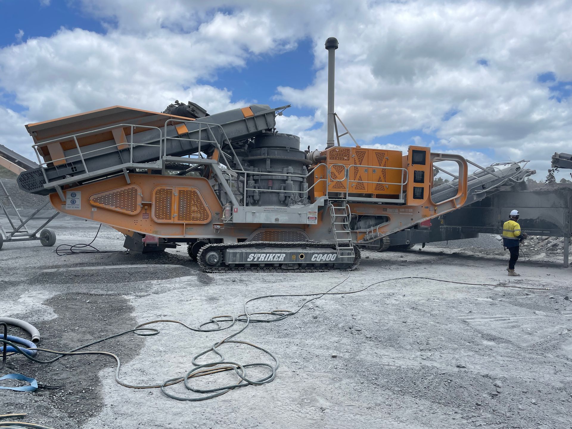 An orange and gray mobile rock crusher at a quarry on a cloudy day. A person in safety gear stands nearby.