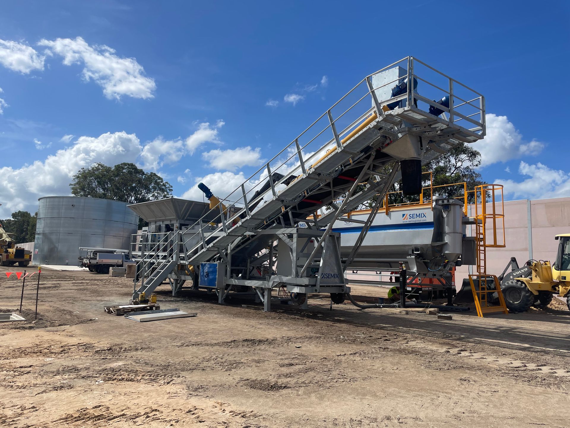An industrial conveyor system on a construction site under a blue sky. A mix of machinery and tanks are in the background.