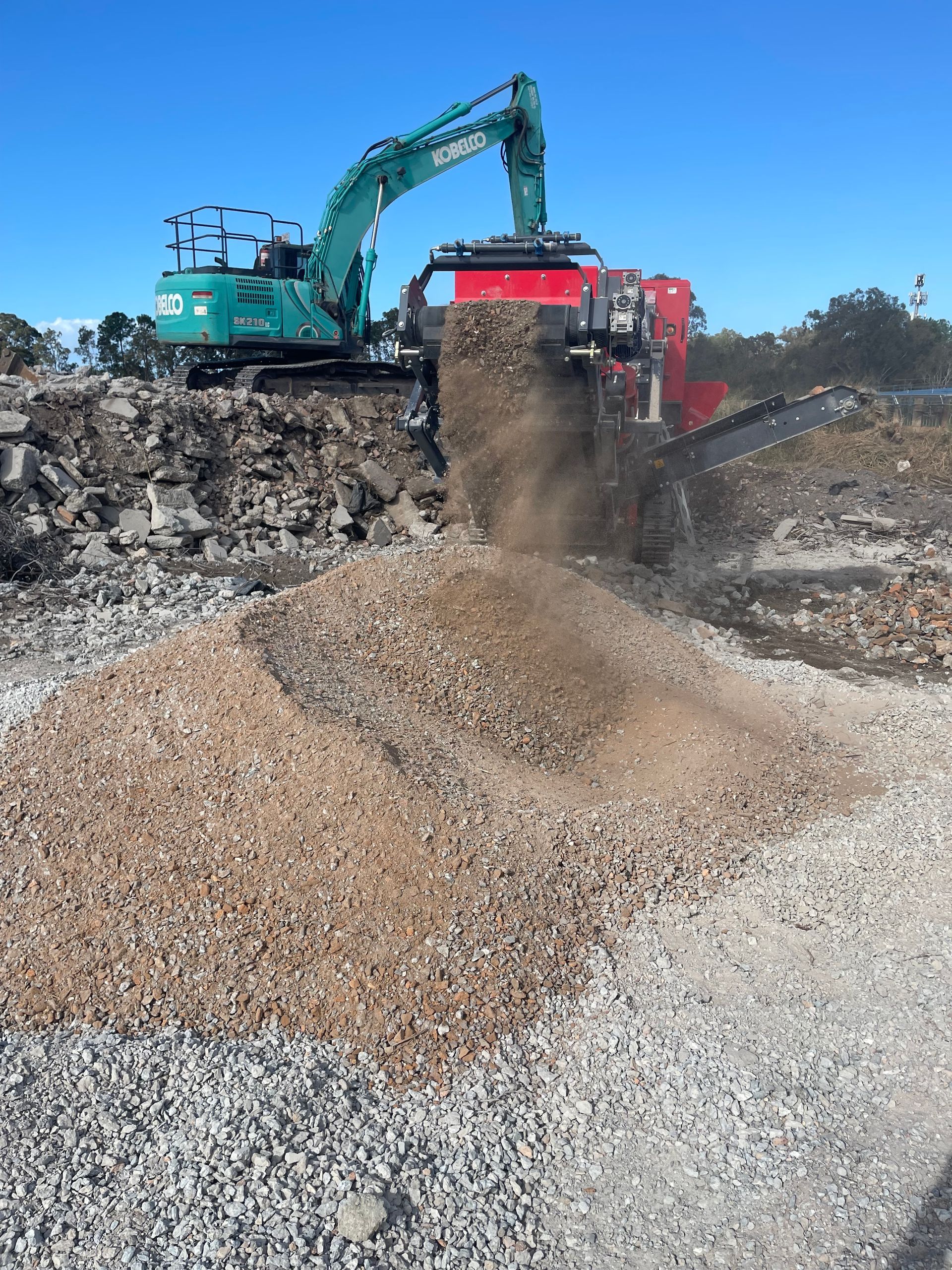 An excavator feeding a mobile crusher, processing demolition debris into gravel on a bright day.