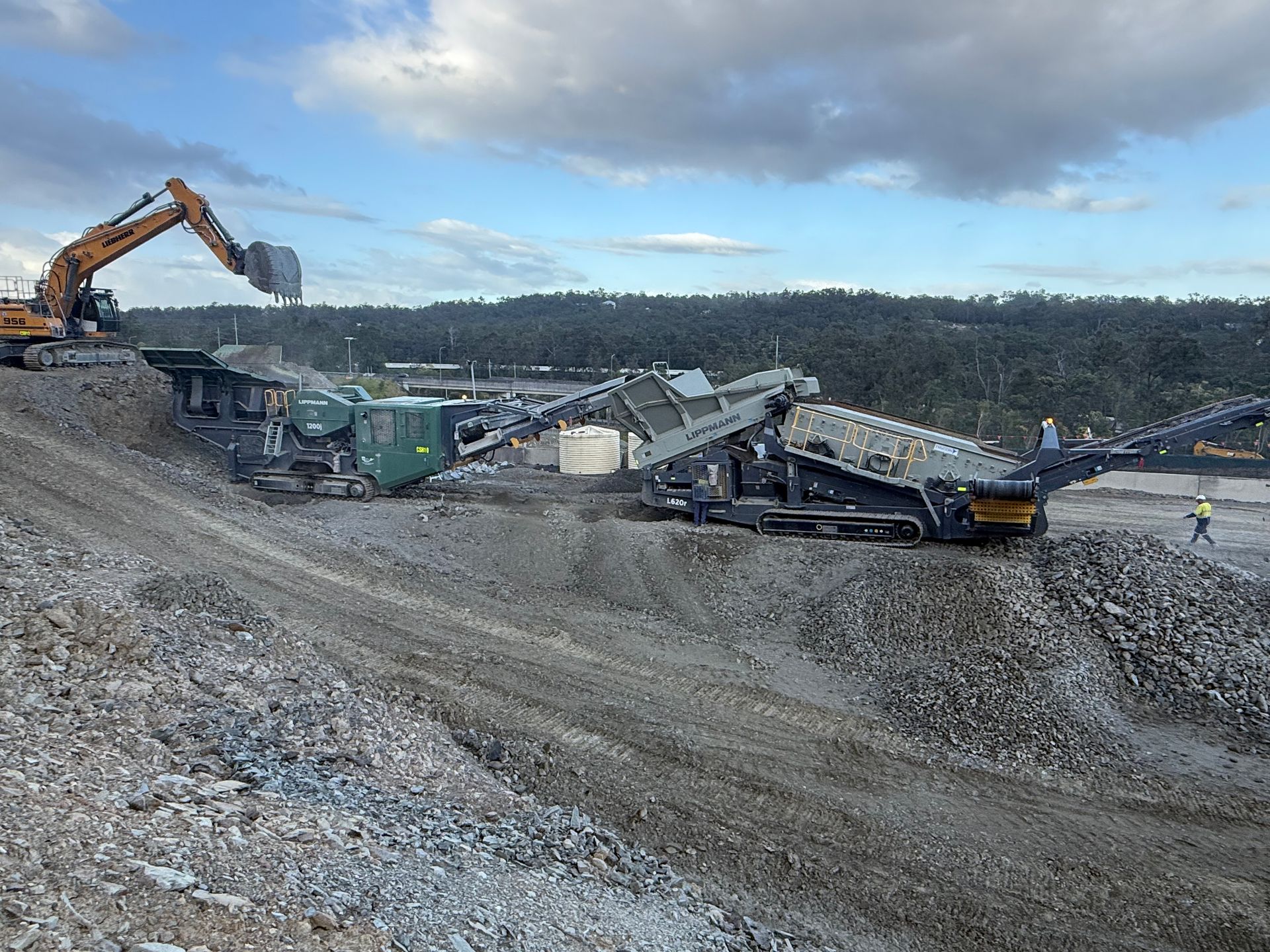 An excavator dumping rock into a mobile processing plant on a pile of rock. Cloudy sky.