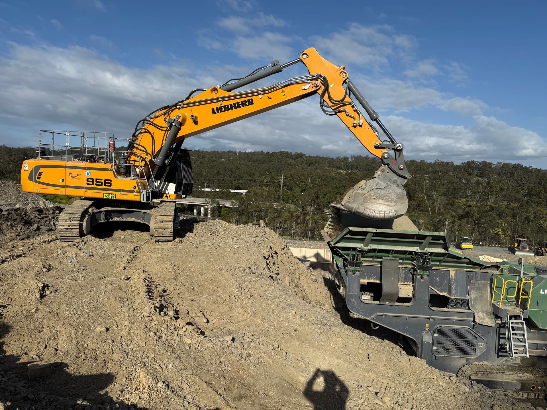 Yellow excavator loading gravel into a screening machine on a dirt pile. Sunny day, blue sky.