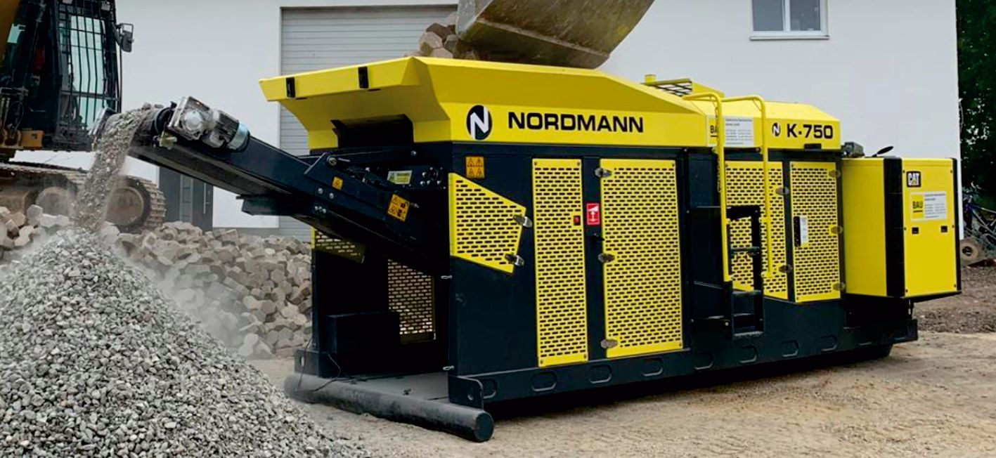 A yellow and black NOROMANN CK 753 machine processing gravel. A pile of gravel is visible near the machine's conveyor belt.