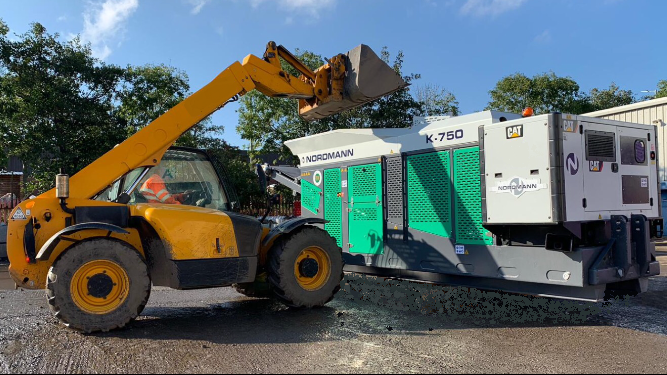 A yellow telehandler loads material into a green and gray industrial machine outdoors.