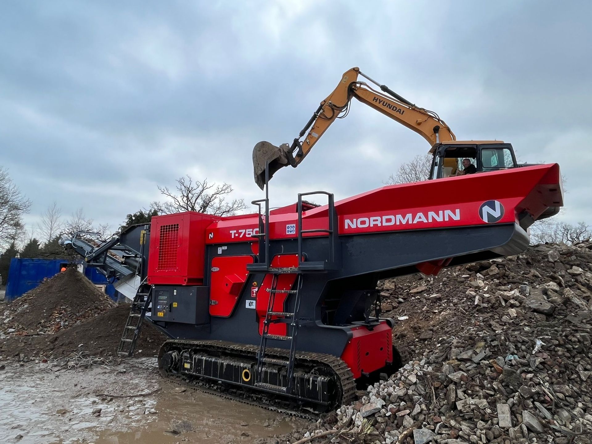 A red Nordmann T-750 shredder on tracks, processing debris with an excavator in a demolition site.