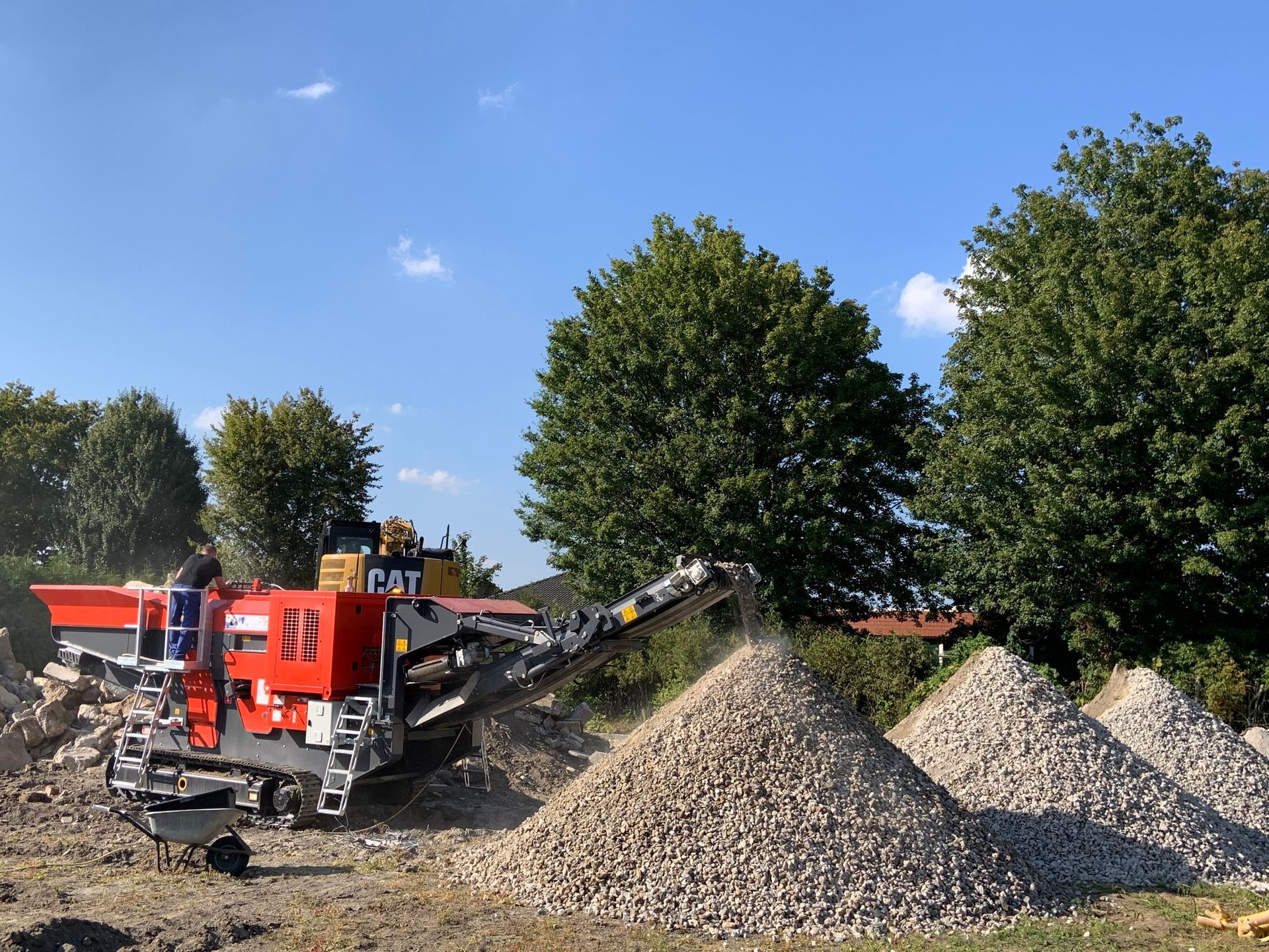 A red and black rock crusher working outdoors, producing piles of crushed stone under a blue sky.