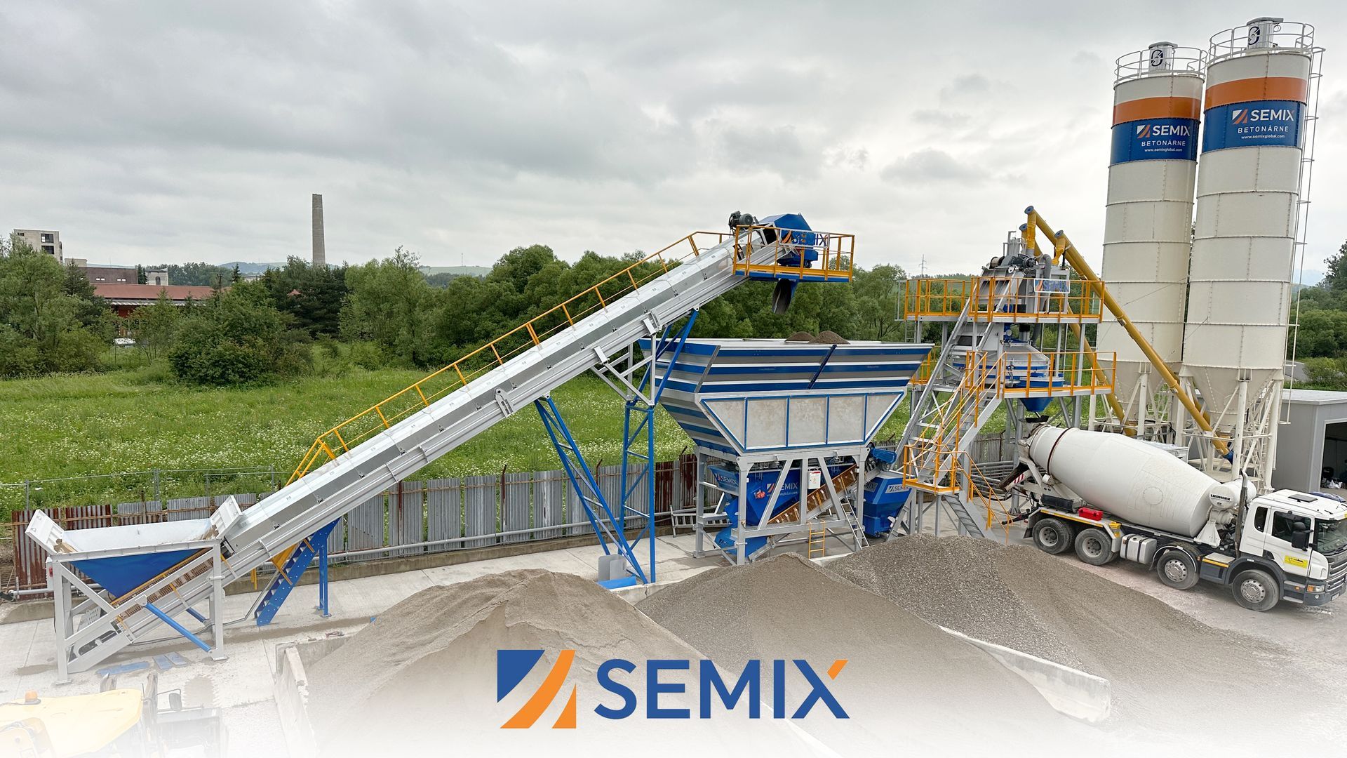 A concrete batching plant with a conveyor belt, silos, and a truck. The plant is blue and white, located outdoors. SEMIX logo is at the bottom.