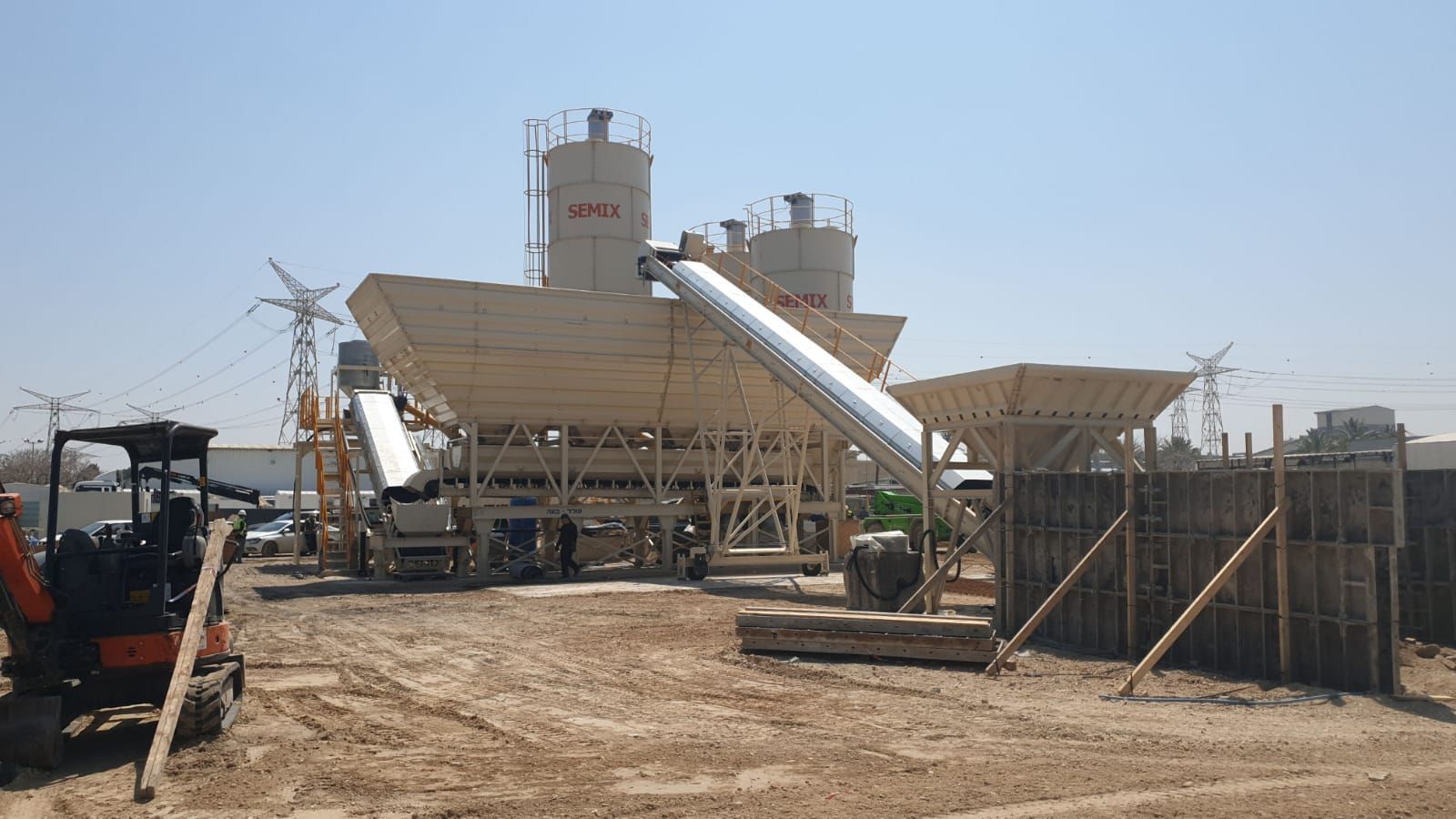 Concrete batching plant with silos, conveyors, and a small excavator on a construction site under a sunny sky.