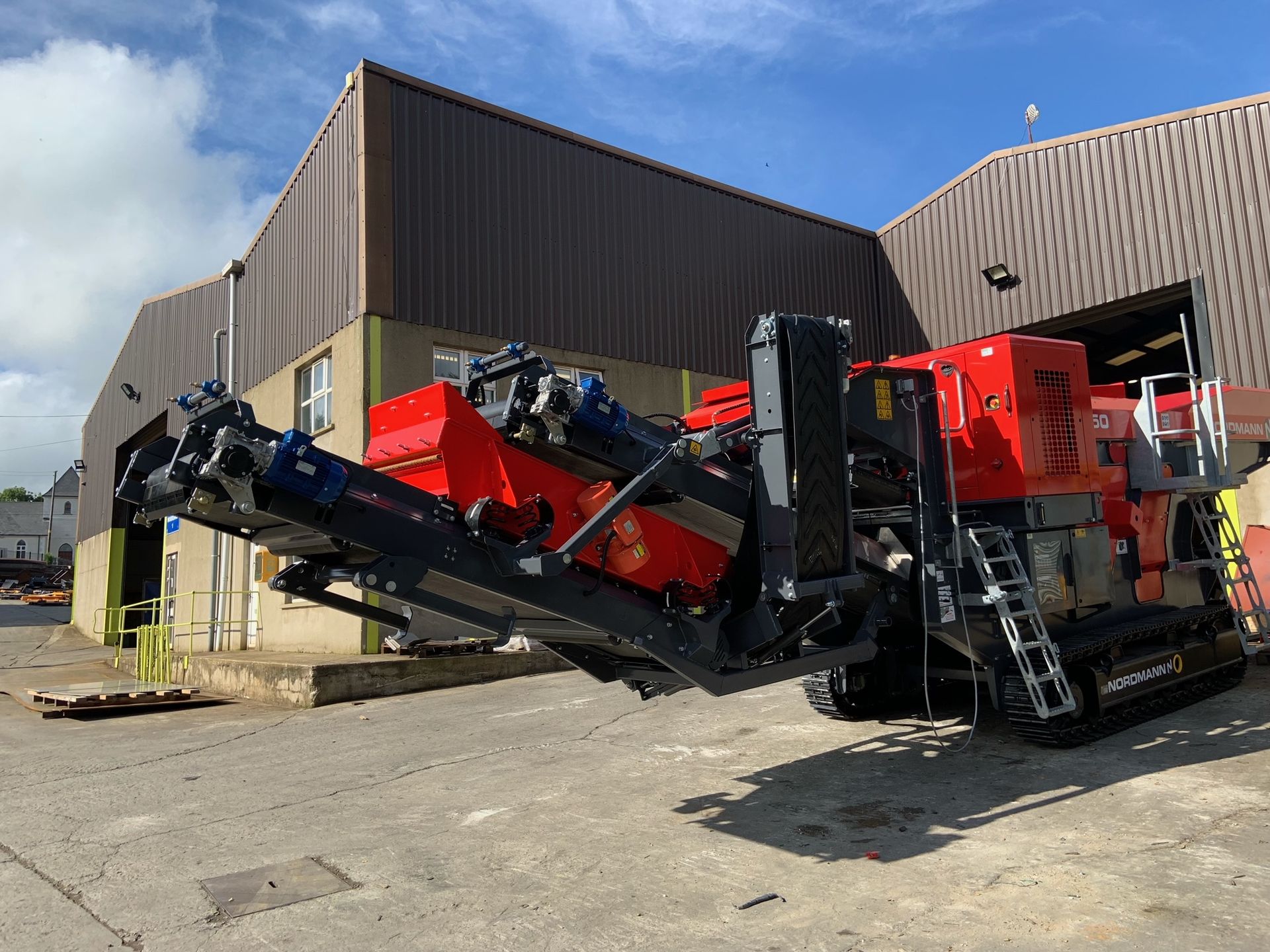 A red and black industrial machine with tracks, likely a rock crusher, sits outside a building.