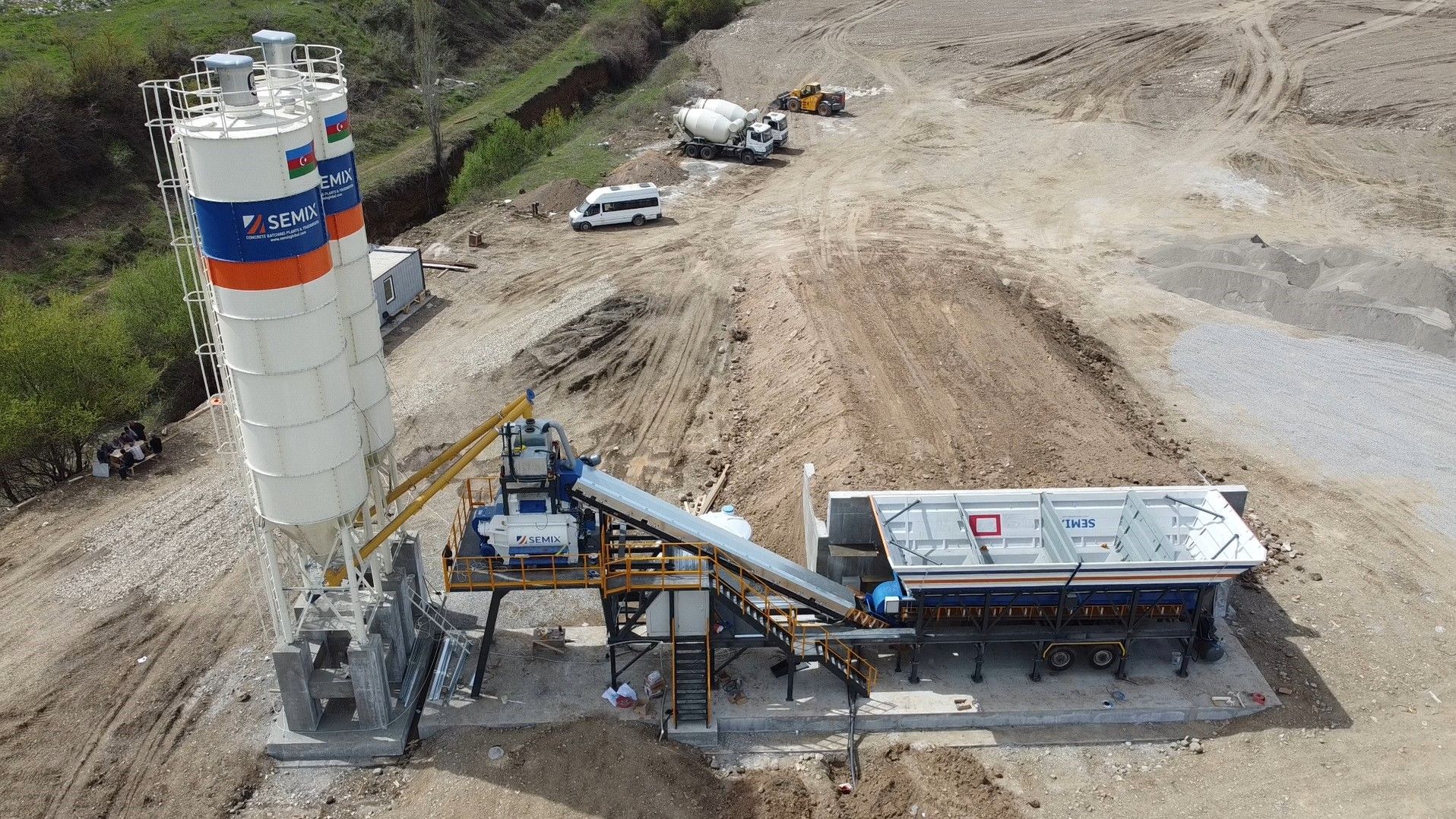 Concrete mixing plant in a construction site, featuring tall silos, conveyor belts, and a mixing unit, with trucks and earth-moving equipment in the background.