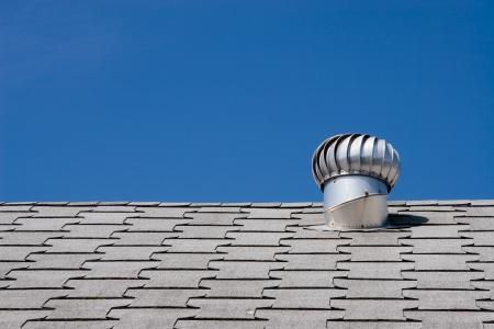 A metal wind turbine roof ventilator mounted on grey asphalt shingles against a clear blue sky.