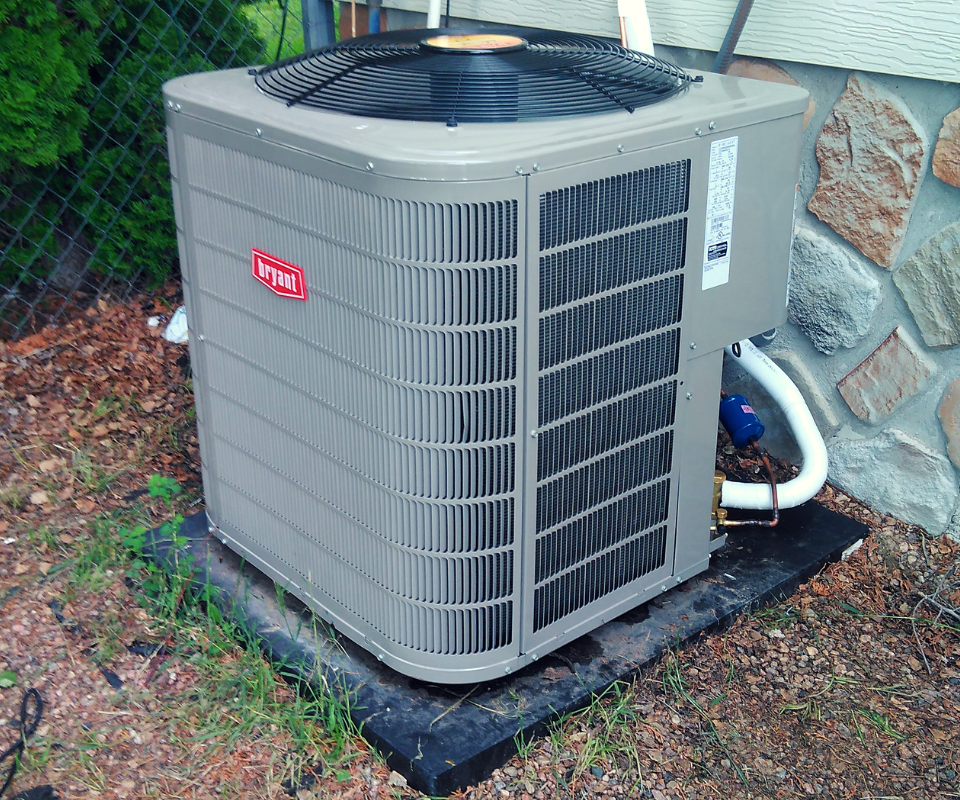 A grey Bryant brand outdoor air conditioning unit sits on a black pad against a stone house exterior.