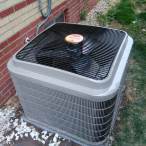 A square, light gray residential air conditioning unit sitting on gravel next to a brick house wall.