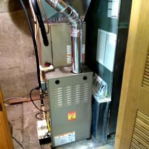 A gray furnace unit with a shiny metal exhaust pipe, situated in a corner near a wooden door frame.