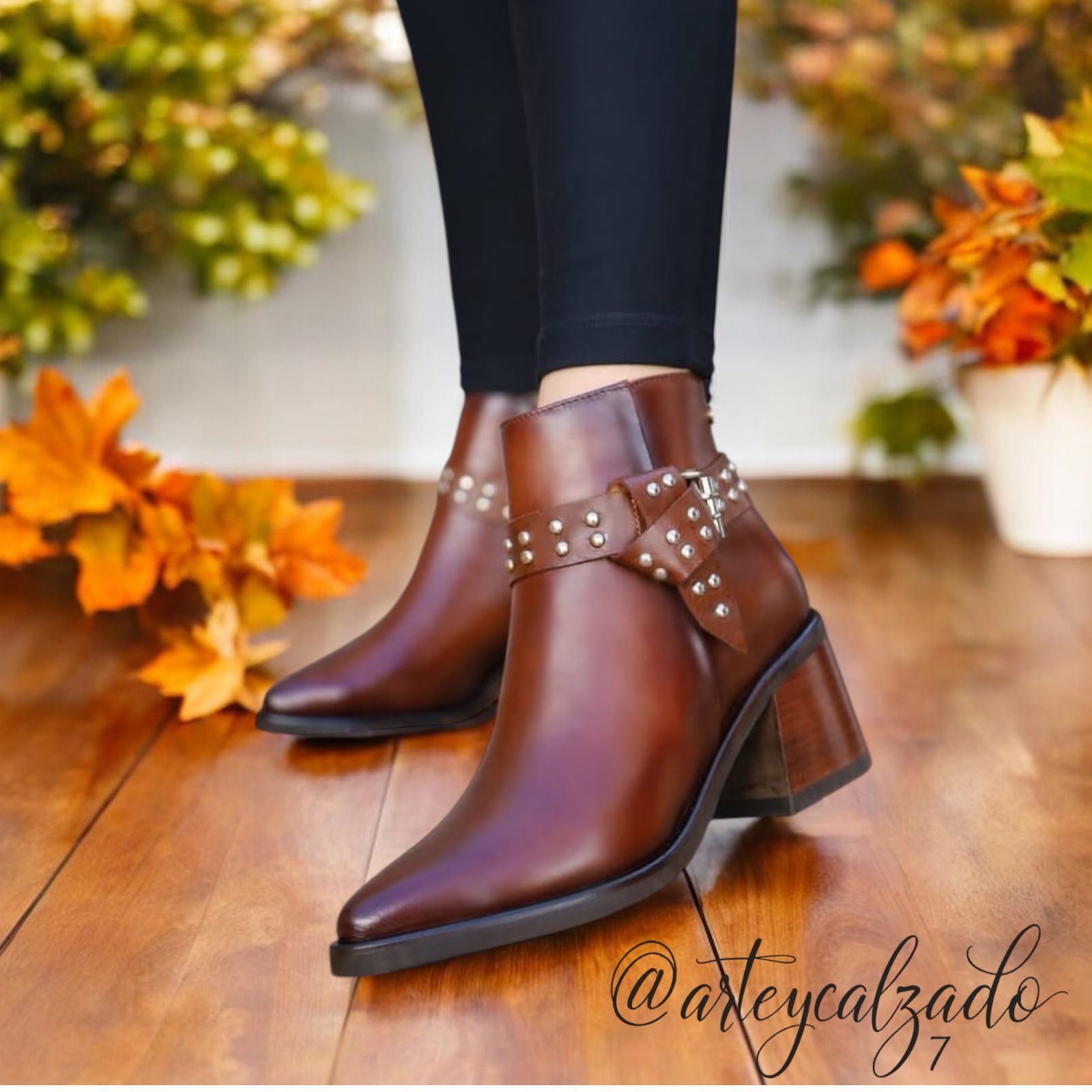Un par de botines de cuero marrón con tiras con tachuelas y tacón de bloque, exhibidos sobre un fondo con temática otoñal.