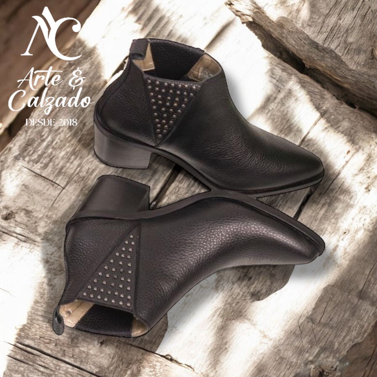 Dos botines de cuero negro con paneles laterales con tachuelas, expuestos sobre una superficie de madera rústica.