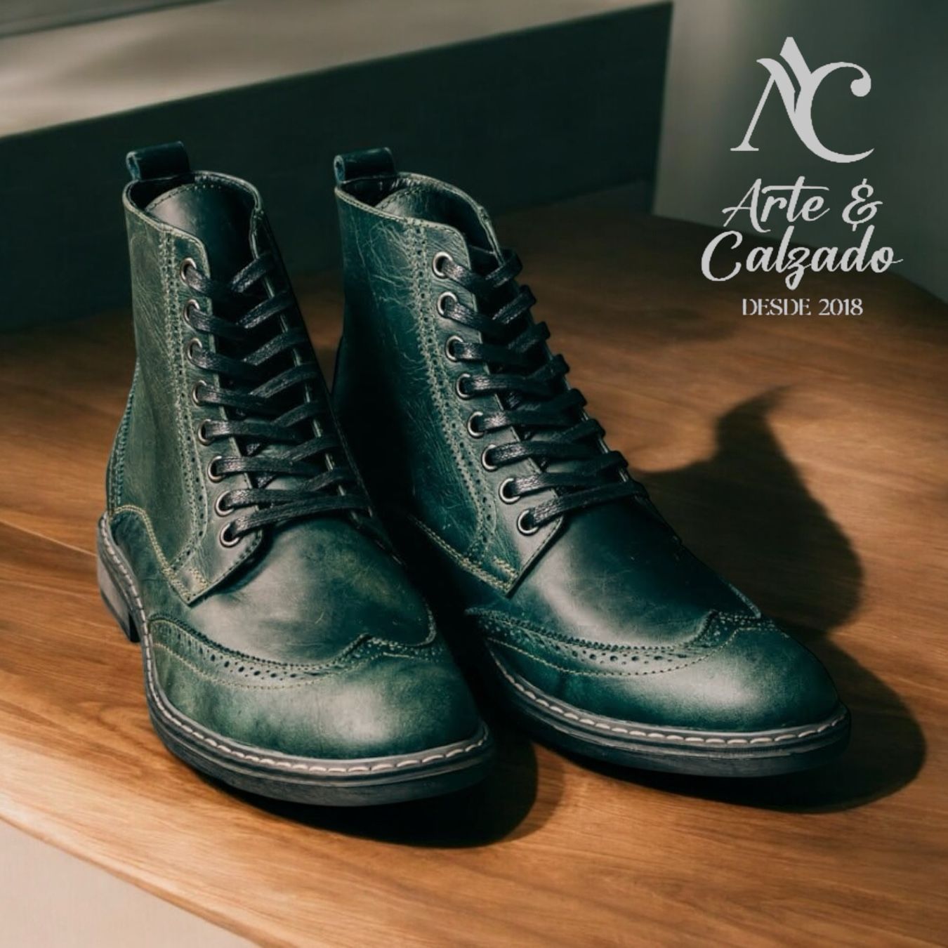 Un par de botas de cuero verde oscuro con puntera perforada y cordones negros sobre una superficie de madera