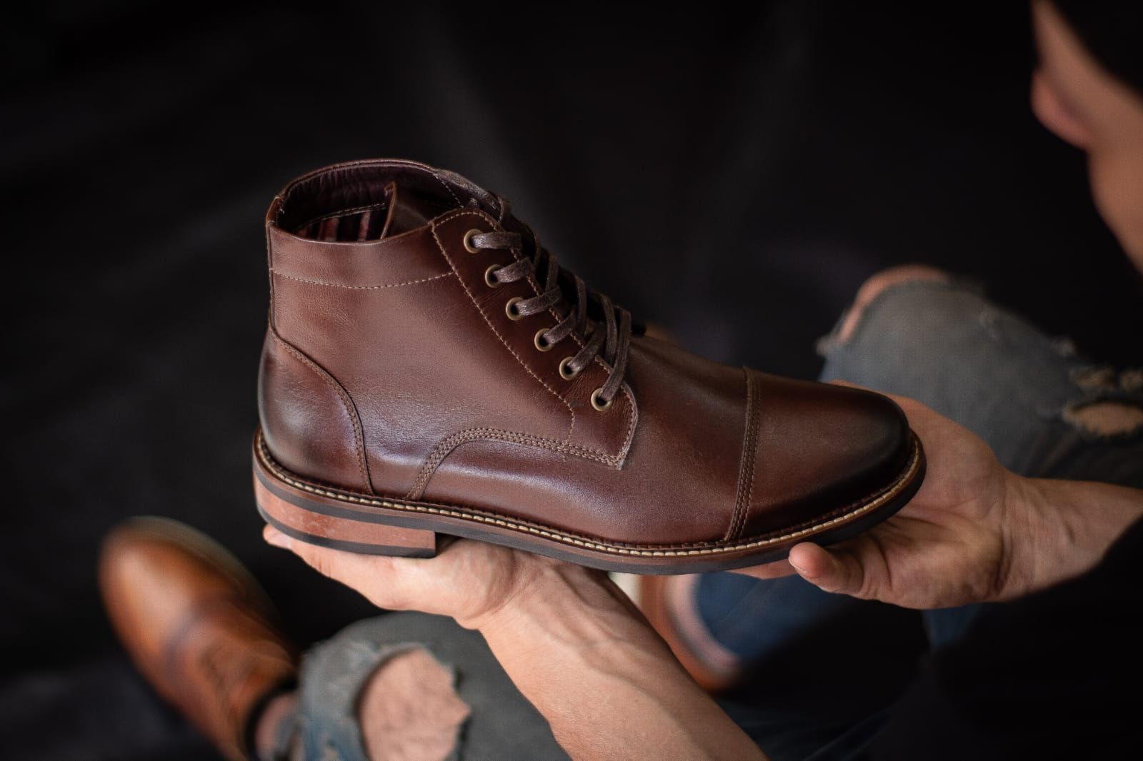 Una persona sostiene una bota de tobillo de cuero marrón con cordones, puntera y talón bruñidos, sobre un fondo oscuro.
