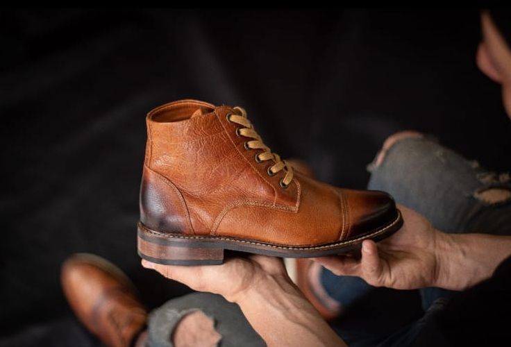 Una persona sostiene una bota de tobillo de cuero marrón con cordones, puntera y talón bruñidos, sobre un fondo oscuro.