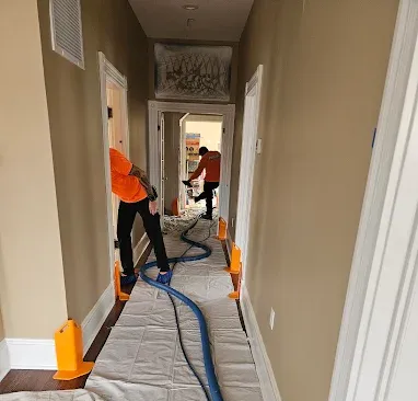 Two people in orange shirts cleaning a hallway with a hose, white drop cloth on floor.