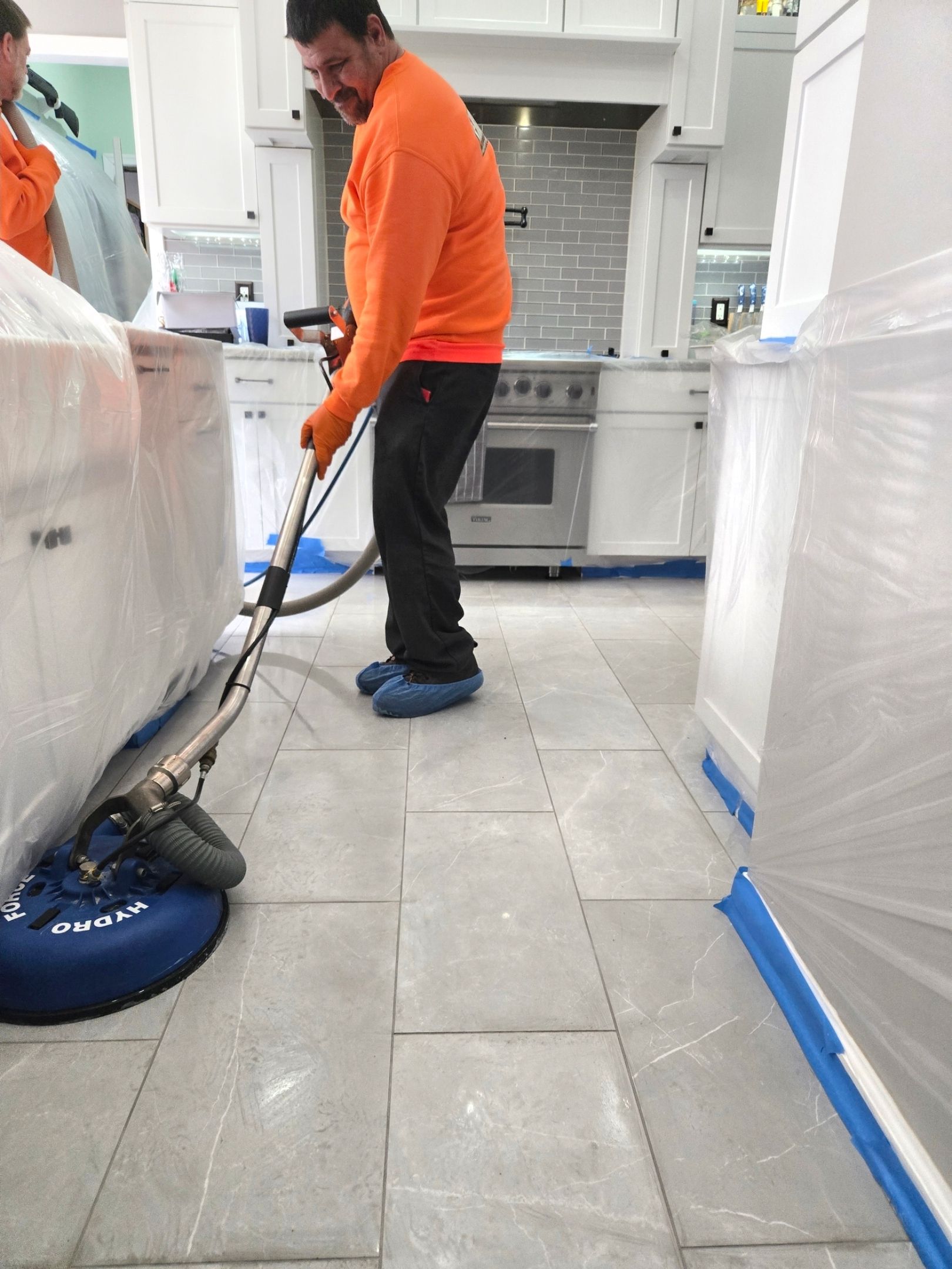 Person in orange shirt cleaning tiled floor in a kitchen with a machine; protected cabinets.