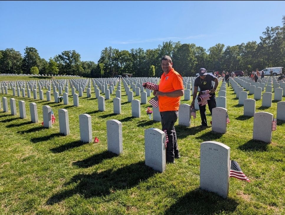 People placing flags at gravestones in a sunny cemetery; commemorating fallen soldiers.