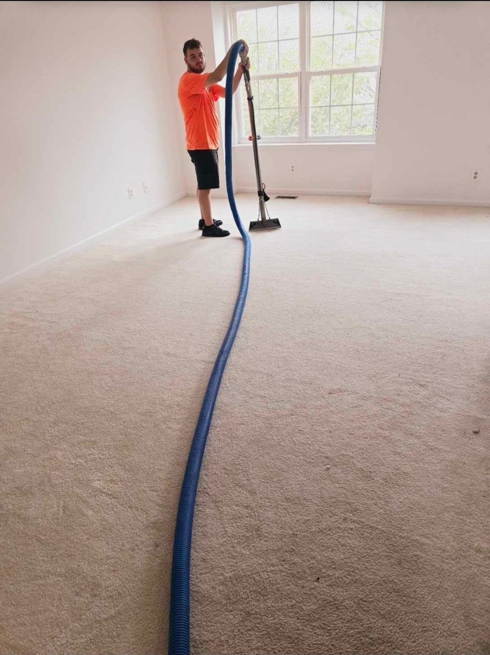 Person in orange shirt cleaning carpet with a vacuum in a room with a window.