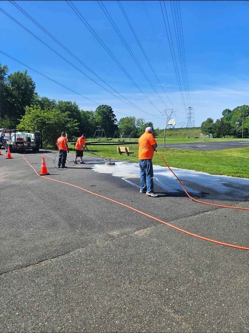 Three workers in orange shirts cleaning a parking lot with a hose. Overhead power lines.