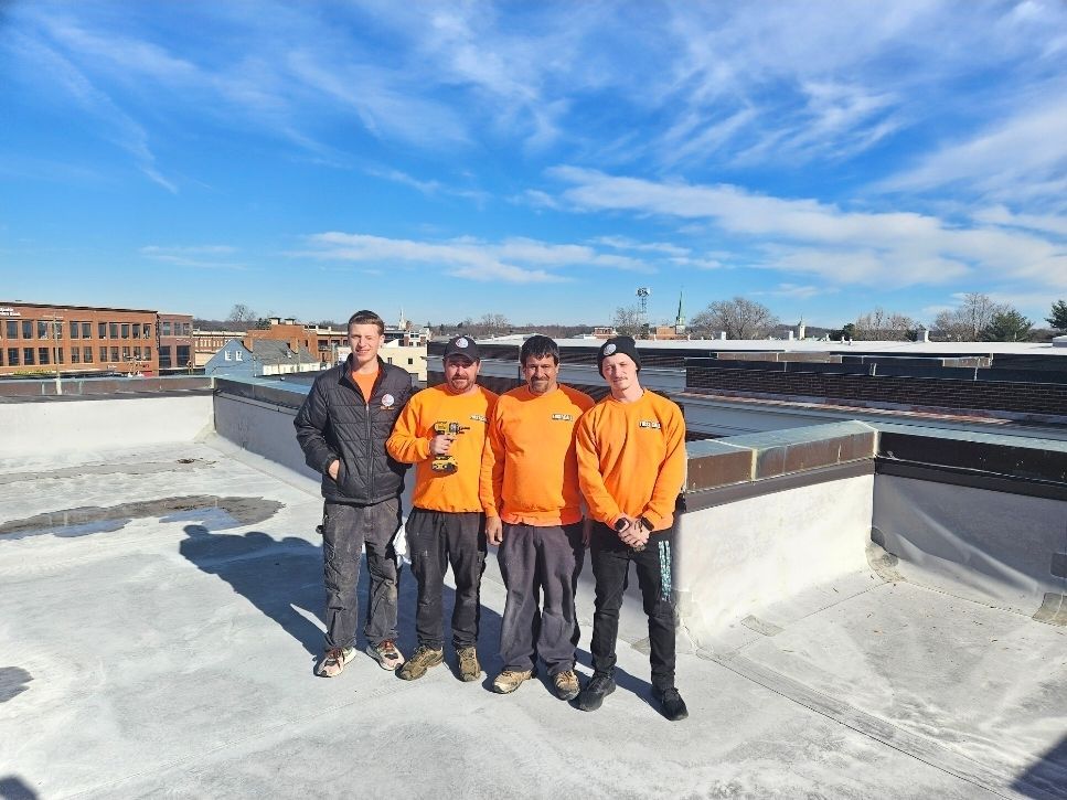 Four workers in orange shirts and a black jacket stand on a flat rooftop on a sunny day.