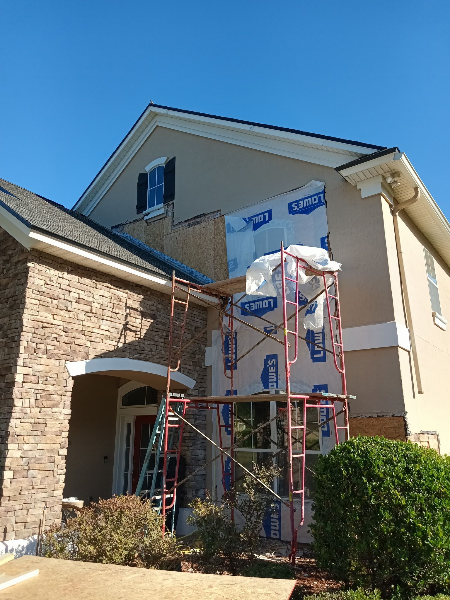 House exterior under construction; scaffolding, blue wrap, beige and stone siding, clear sky.