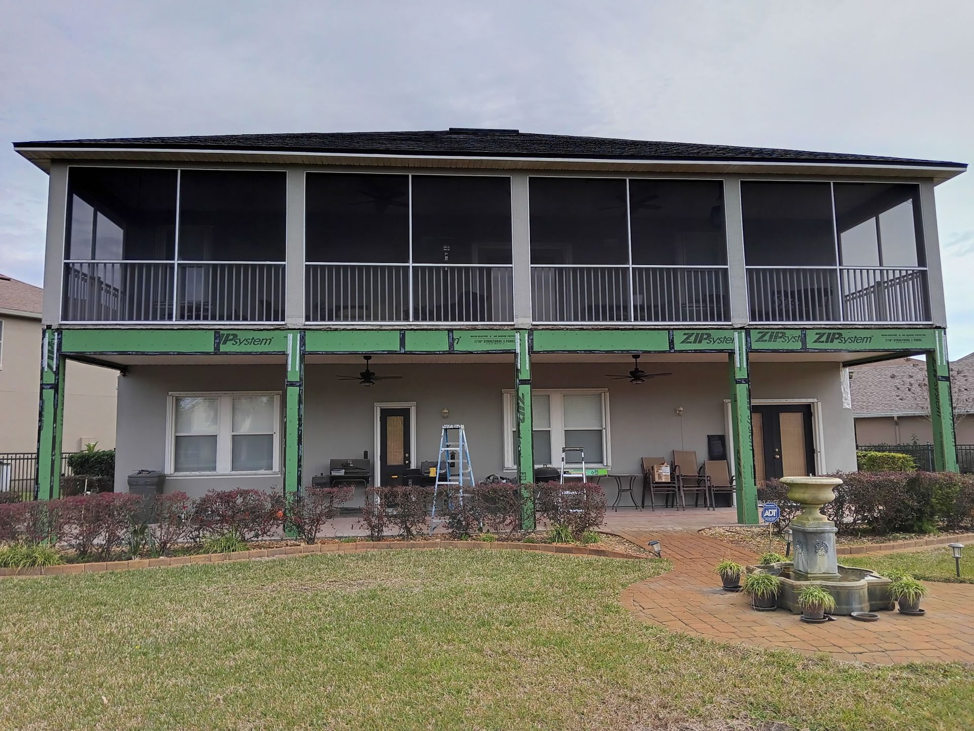 Two-story house with screened porch. Green beams support second-story, backyard with roses and fountain. Cloudy sky.
