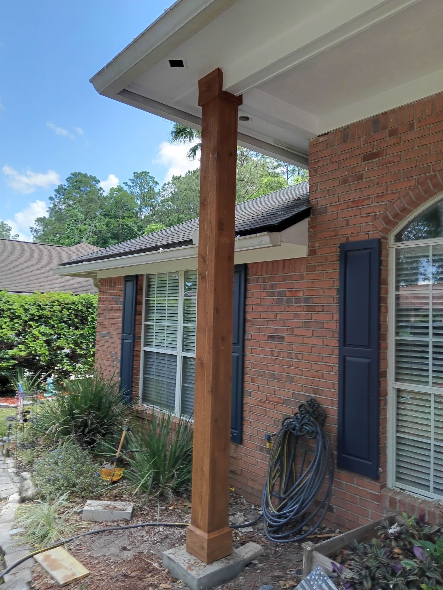 Brown wooden porch column on a brick house. Blue shutters and lush greenery surround the entrance.