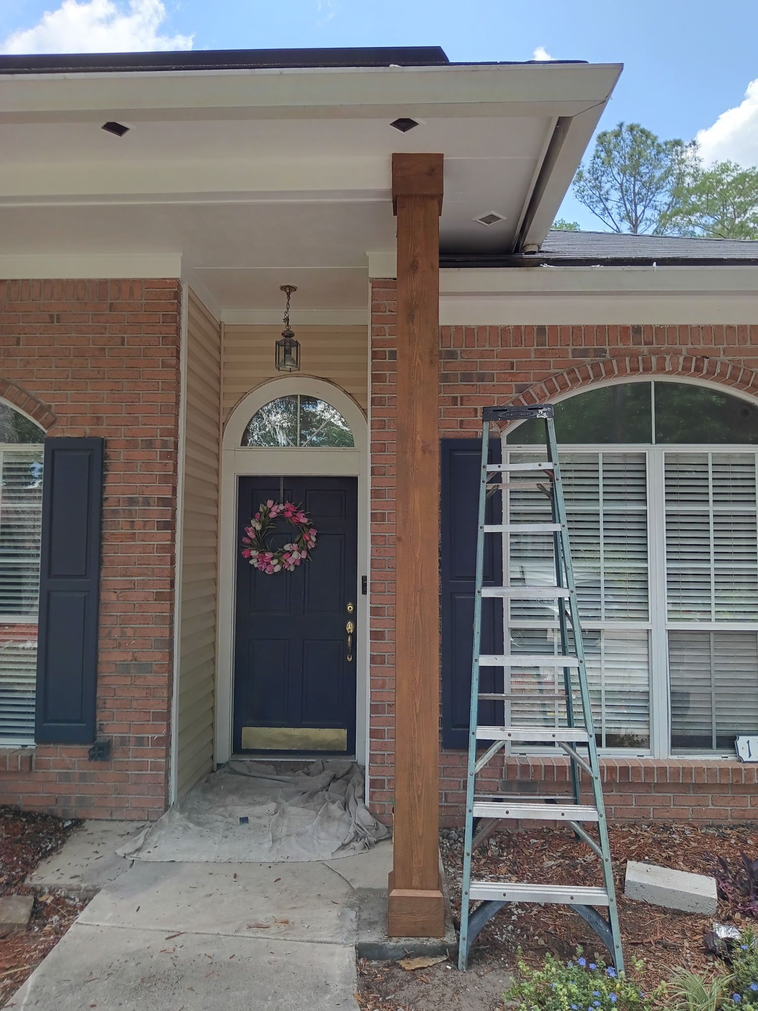 A wooden post supporting a porch roof, ladder beside it. Red brick house with dark blue door and shutters.