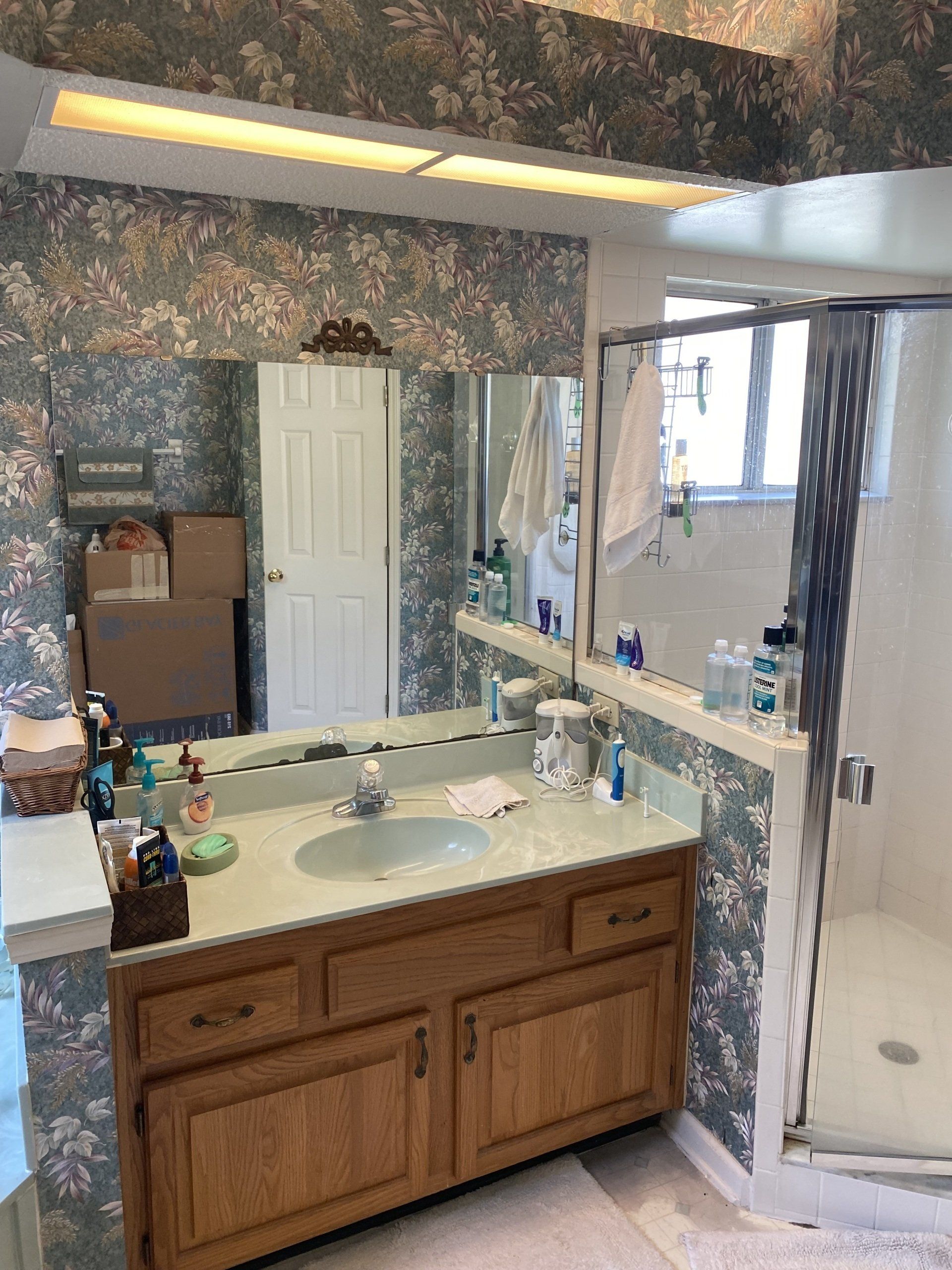 Bathroom with floral wallpaper, wood vanity, sink, mirror, and shower.