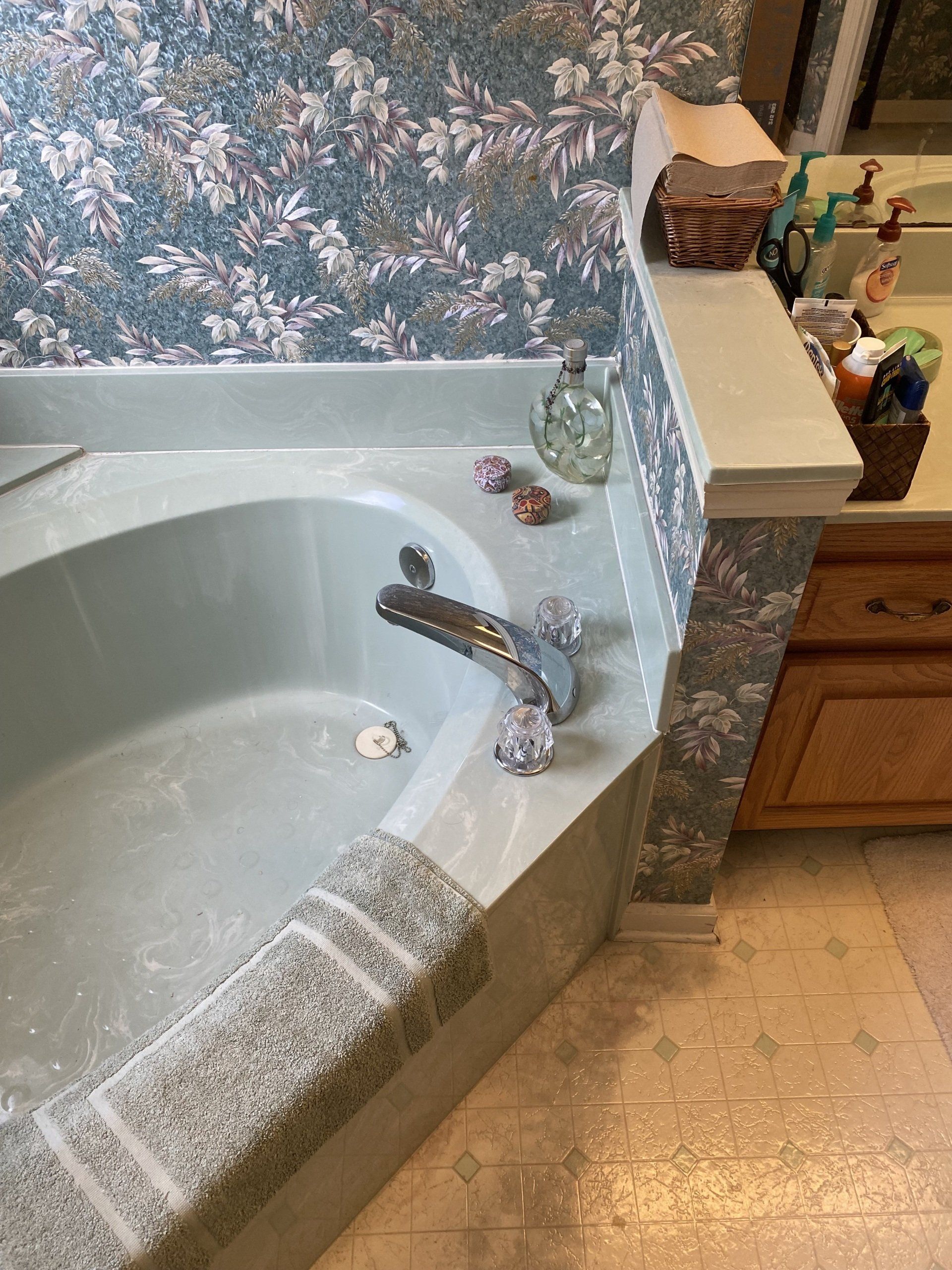 Corner bathtub in bathroom with floral wallpaper and pale green countertop.