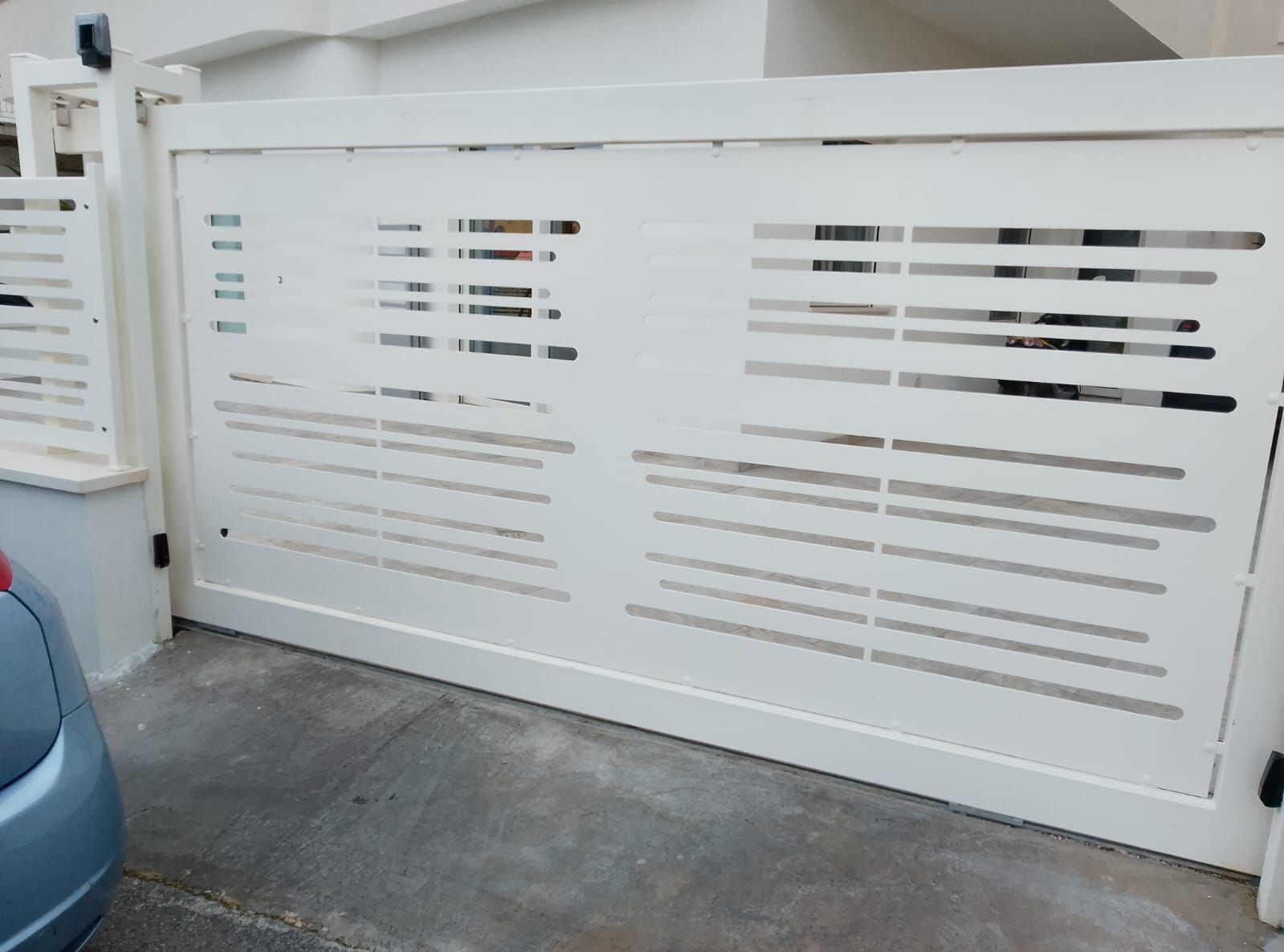 Un grande cancello scorrevole orizzontale bianco con lamelle rettangolari strette, situato su un vialetto di accesso a un'abitazione privata.