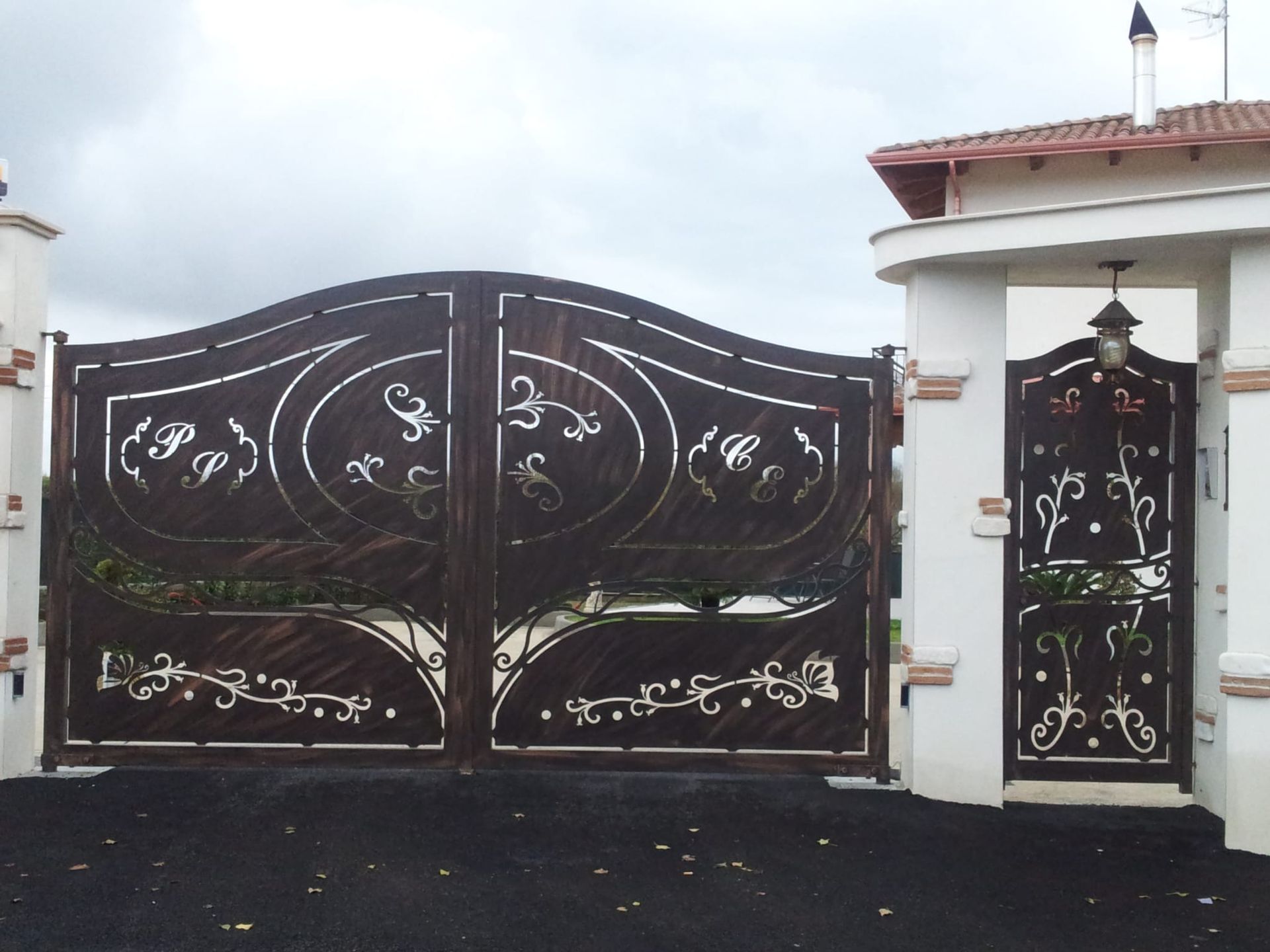 Doppio cancello carrabile in metallo marrone scuro e decorato, con un cancello pedonale abbinato, inserito in un ingresso intonacato di bianco.