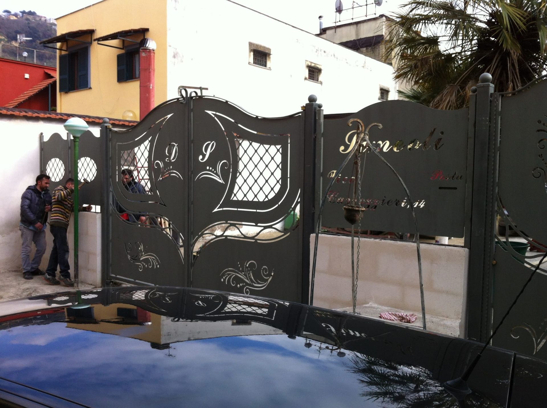 Due persone sono in piedi accanto a un cancello di metallo scuro con intagli decorativi, viste da sopra il tetto lucido di un'auto.