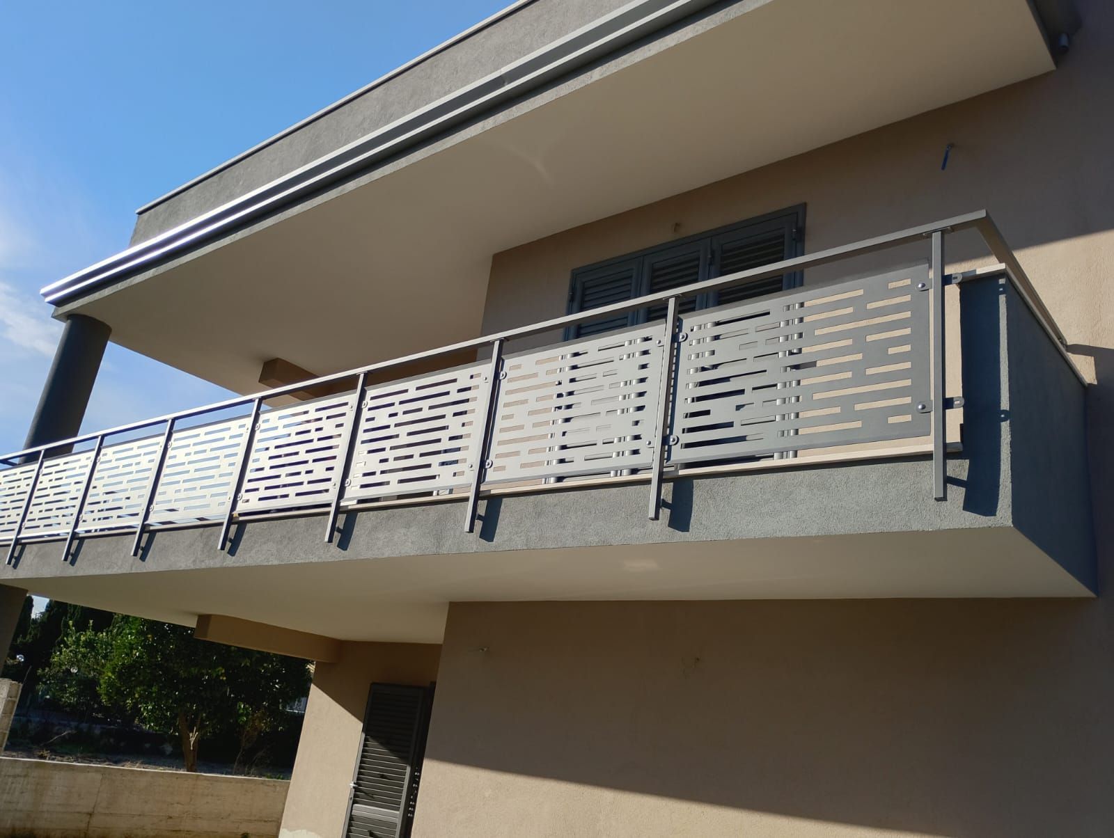 Un balcone con una ringhiera moderna composta da pannelli metallici grigi con fessure orizzontali, fissati a montanti verticali scuri.