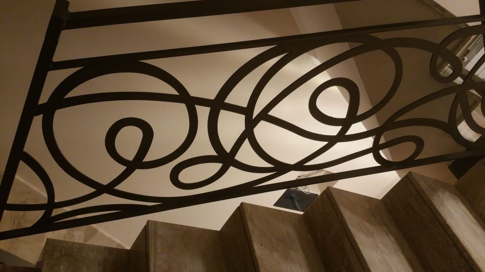 Ringhiere metalliche scure e sinuose su una scala, illuminate da una luce soffusa proveniente dall'alto.